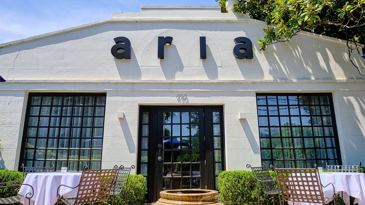 The exterior front entrance of Aria with simple lettering over the door and large windows plus tables with white cloths and brown chairs in Atlanta, Georgia, USA